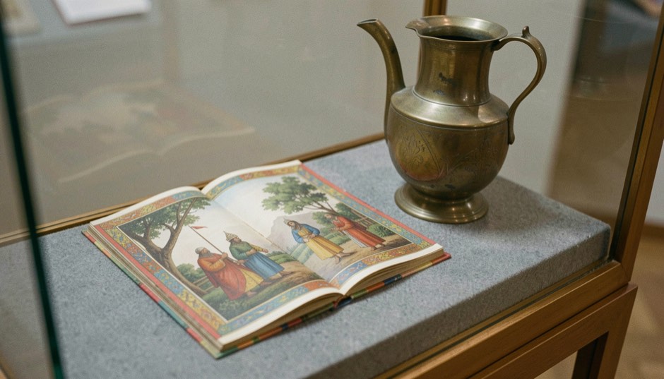 In einer Museumsvitrine liegt eine aufgeschlagene Mogulminiatur mit farbigem Rand neben einem alten Messinggefäß auf grauem Filz.